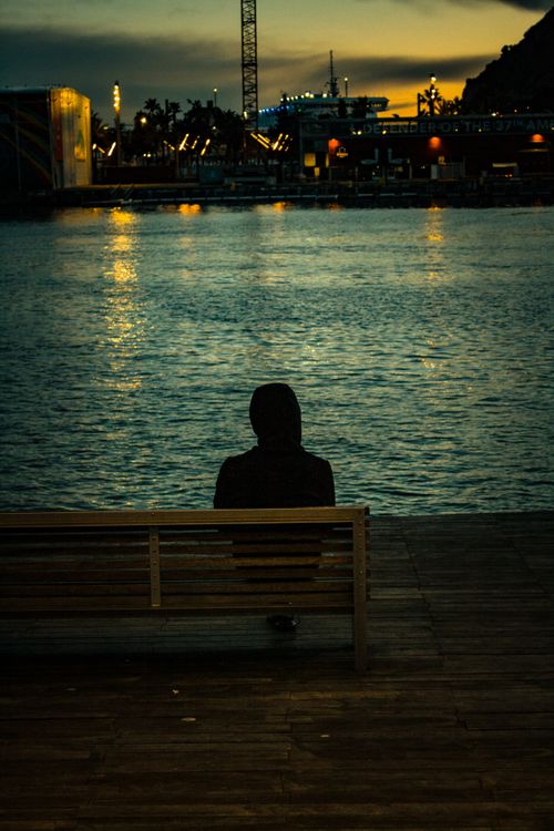 Free Person in hoodie seated on a bench by water during twilight, facing city lights. Stock Photo