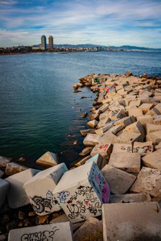 Concrete breakwaters with colorful graffiti line the coast of Barcelona, with city skyline in the background.