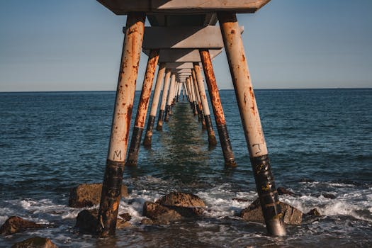 A view under a rusty pier extending into the ocean with waves crashing against rocks.