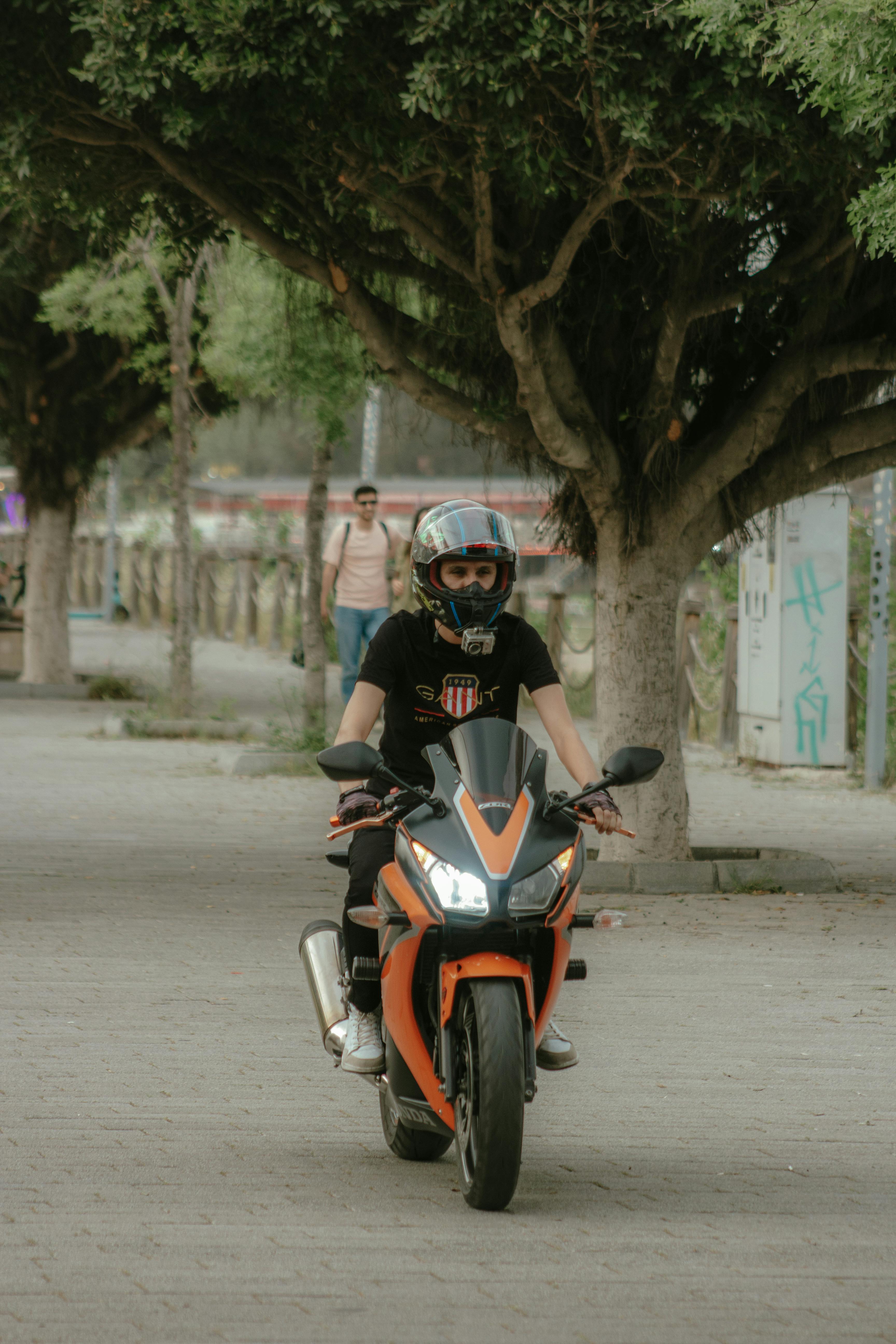 A motorcyclist wears a helmet while riding on a sidewalk lined with lush trees in an urban setting.