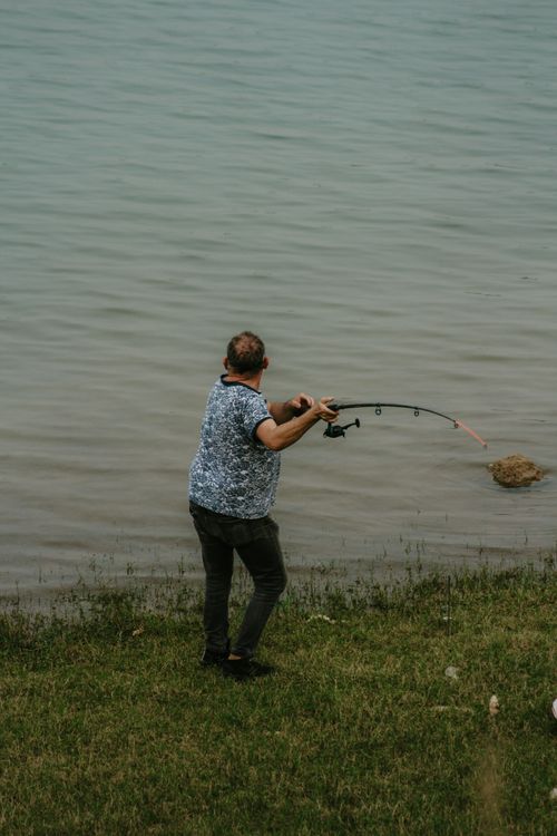 Free A balding man stands by the lakeshore holding a fishing rod, casting into the water. Stock Photo