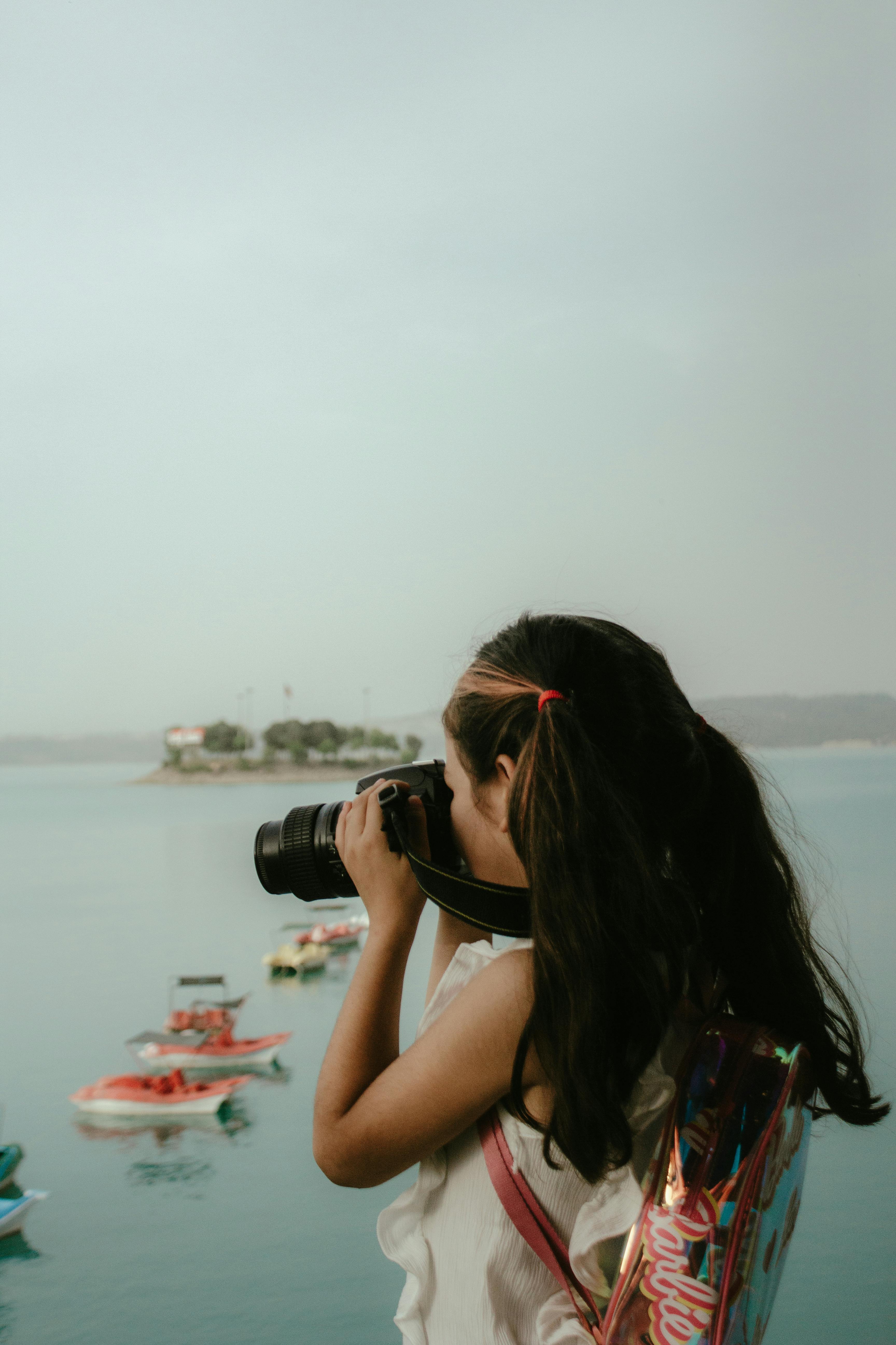 Free A young girl with long hair photographs the serene coastline, focusing on colorful boats. Stock Photo
