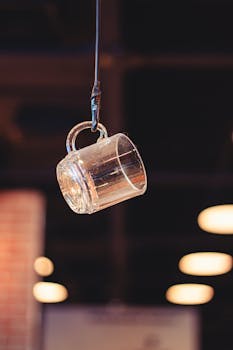 A glass mug hanging from a wire in a warmly lit indoor cafe setting with blurred background.