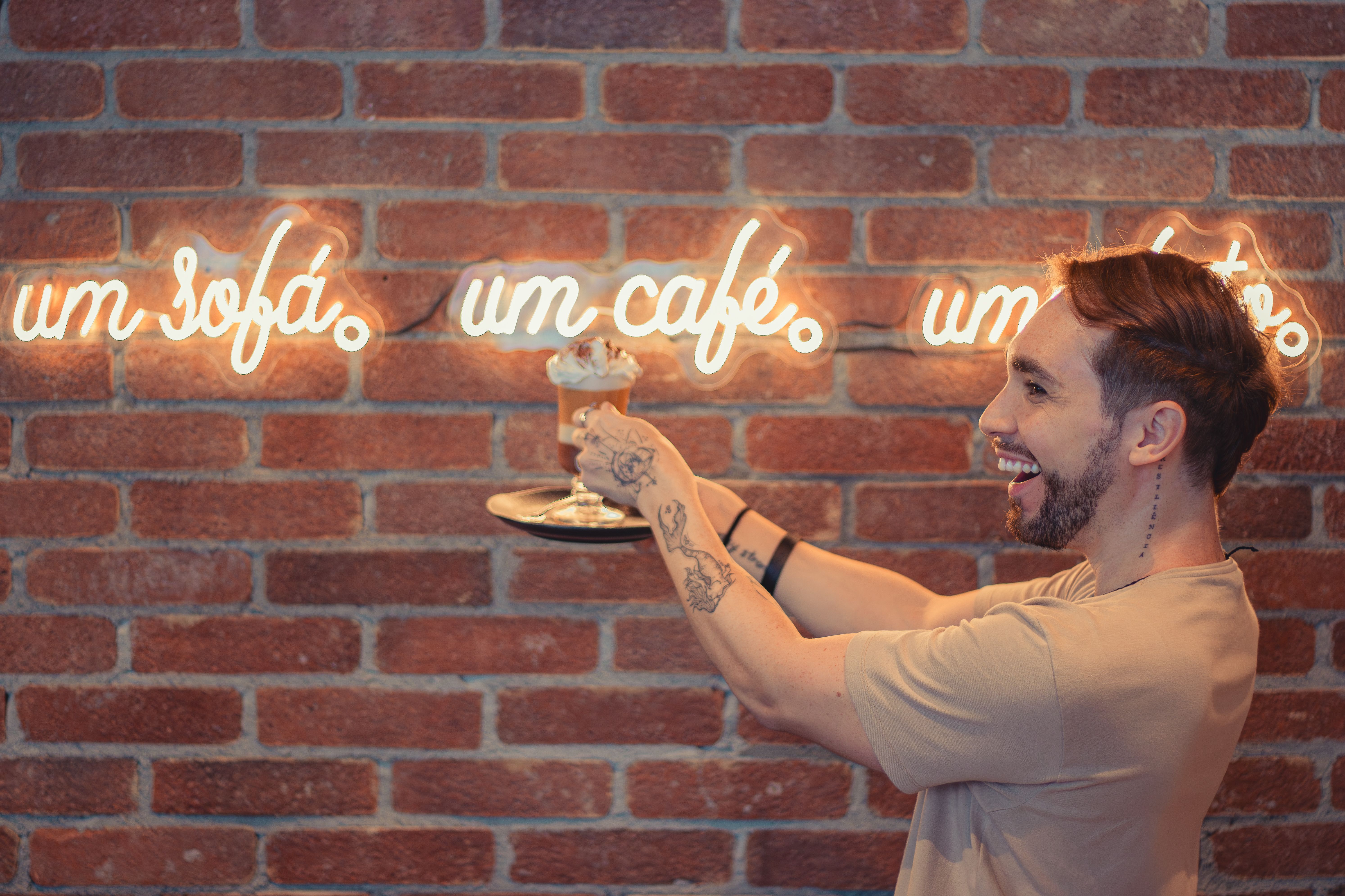 A joyful man with tattoos holds a coffee drink against a neon-lit brick wall in a café.