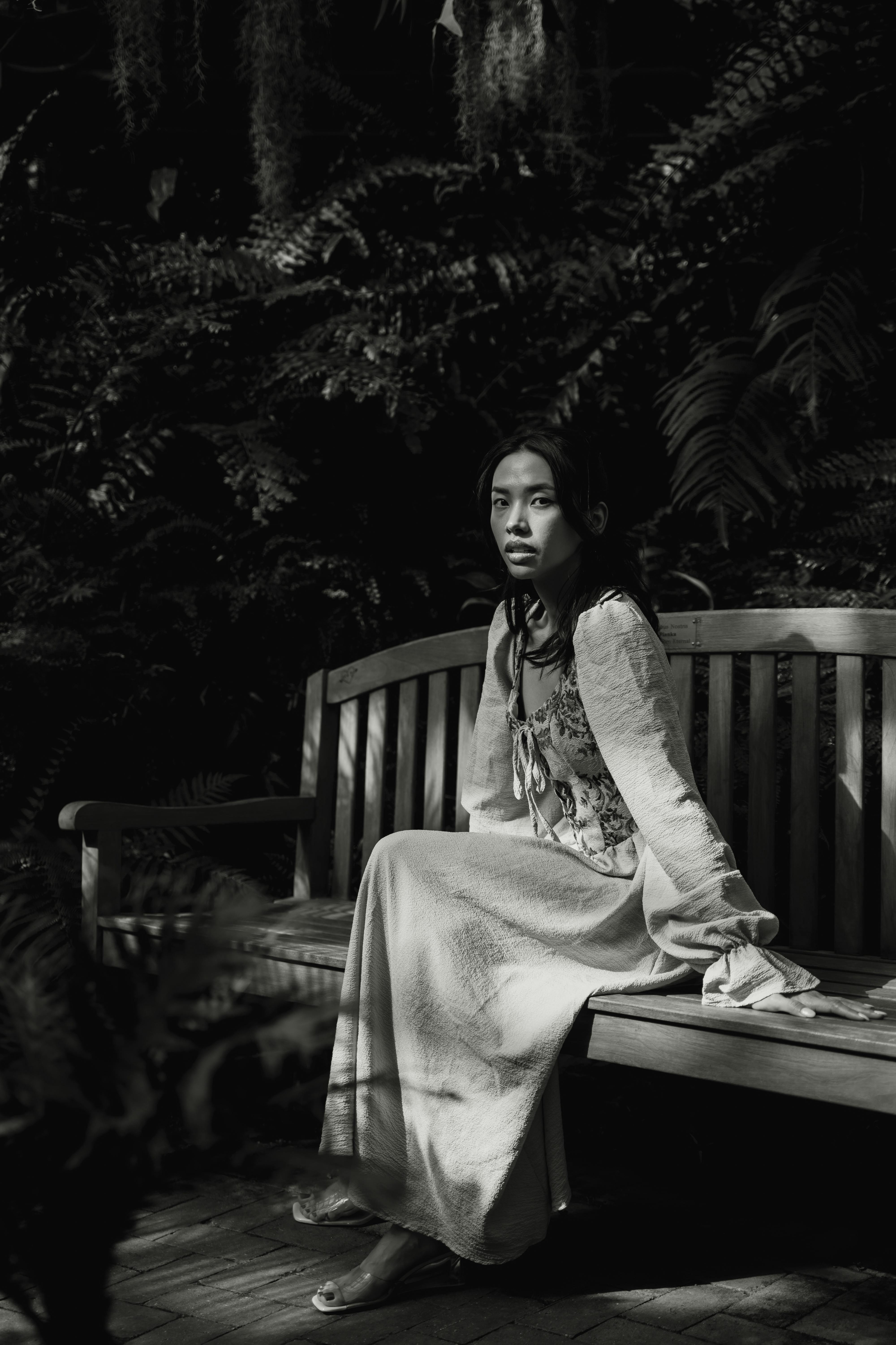 Monochrome photo of a woman in a dress sitting on a wooden bench surrounded by foliage.