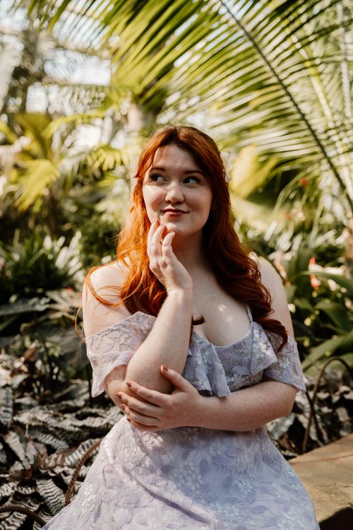 Free A red-haired woman in a lavender dress posing thoughtfully in a lush botanical setting. Stock Photo