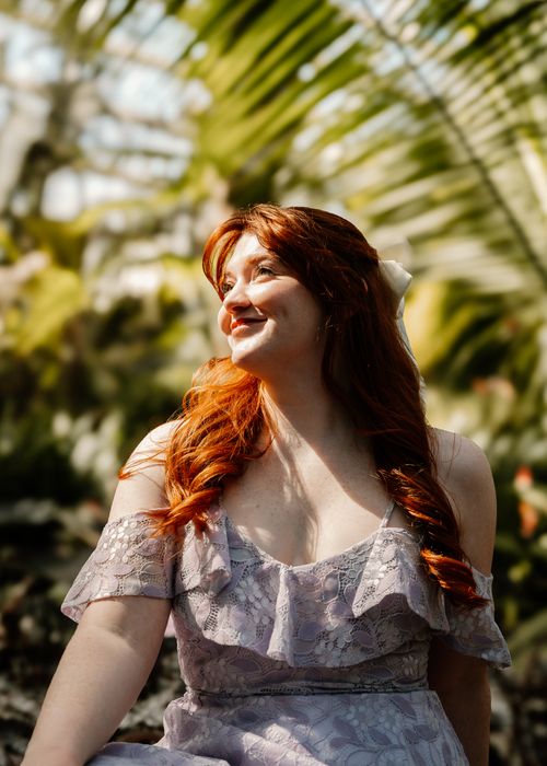 Free A woman in a floral dress enjoys the sunlight in a lush garden setting, surrounded by greenery. Stock Photo