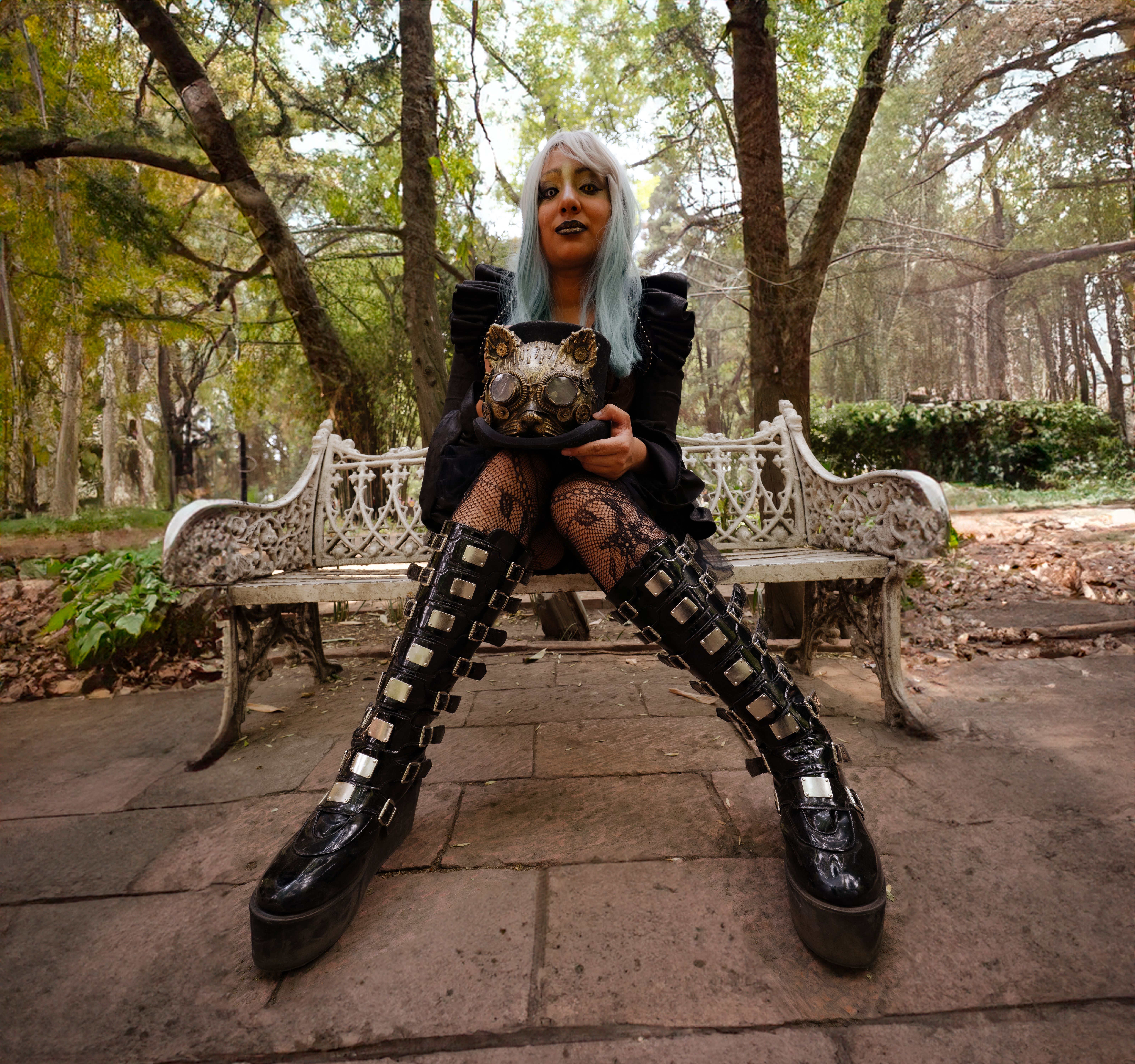 Gothic woman in punk attire holds cat mask on a park bench in Mexico City's leafy setting.