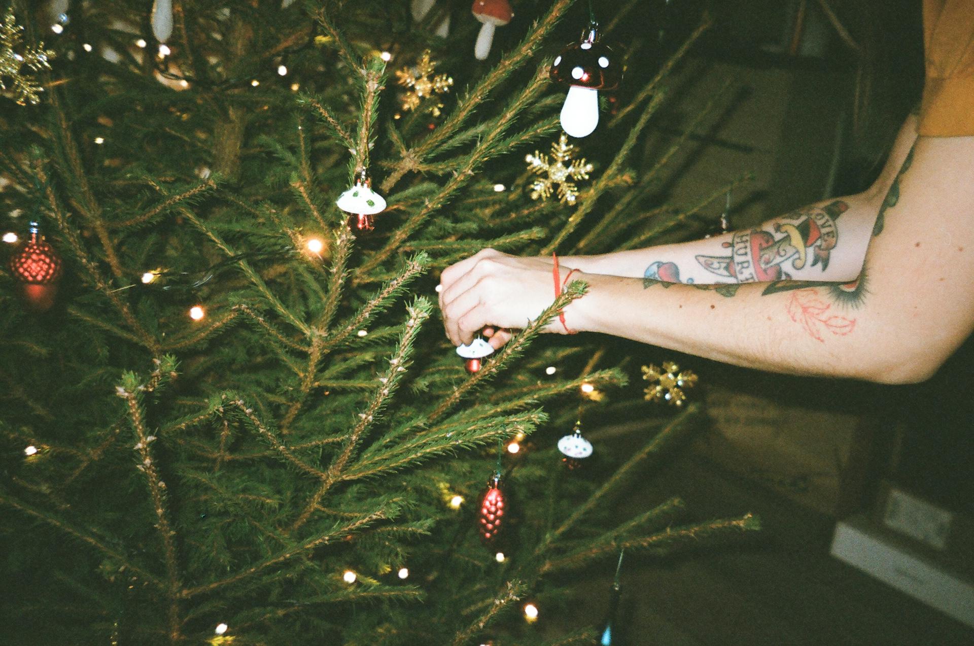 A person decorates a Christmas tree with ornaments featuring unique tattooed arms.