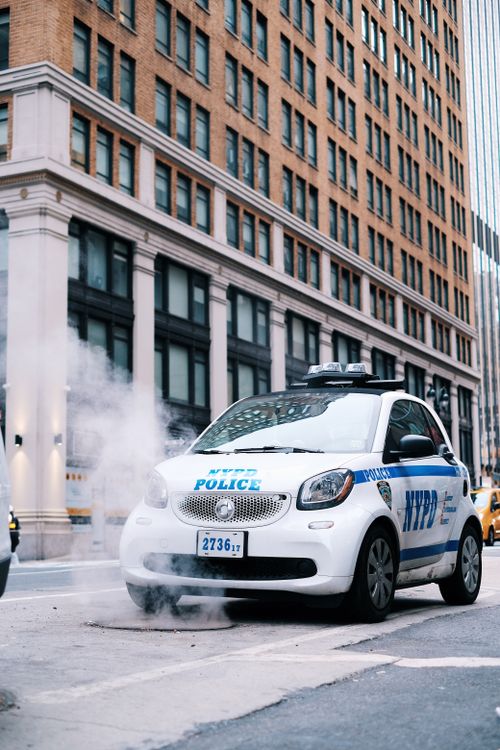 Free A New York police car parked by a city building with steam rising, showing urban life. Stock Photo