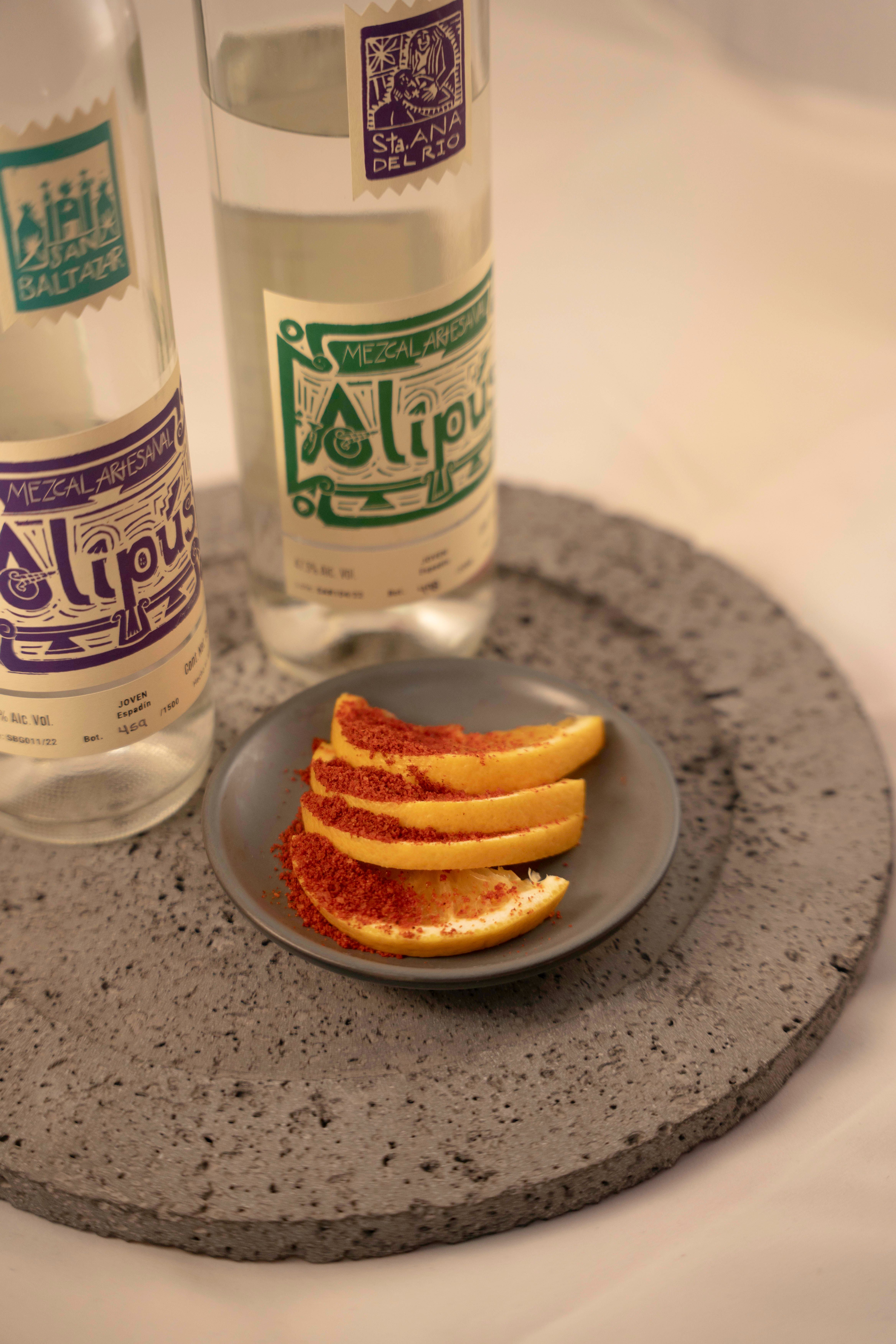 Close-up of mezcal bottles with seasoned orange slices on a textured stone plate, showcasing a traditional Mexican drink.