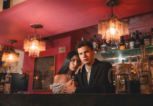 A stylish couple poses at a sophisticated bar, illuminated by warm, ambient lighting and chandeliers.
