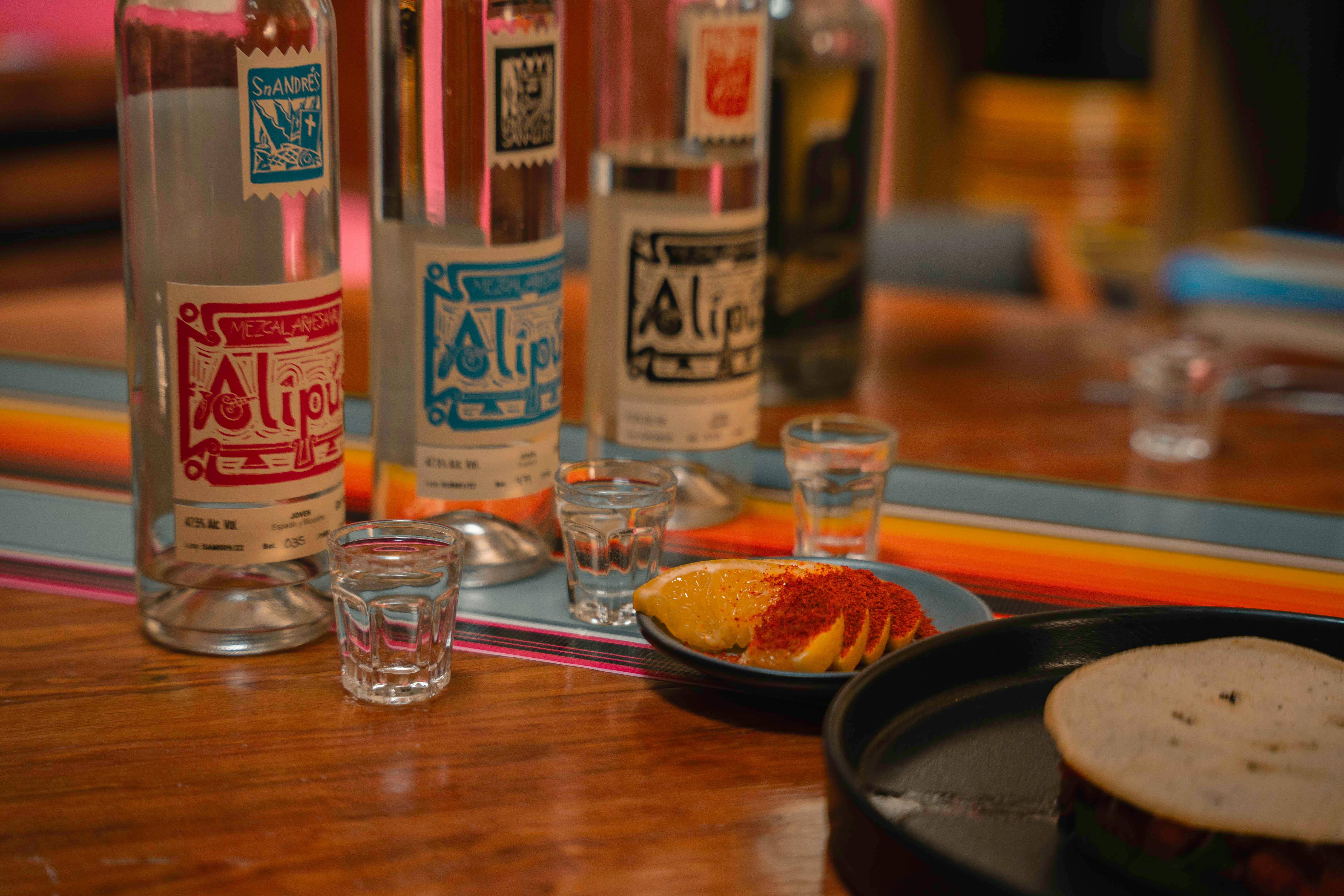 Colorful close-up of mezcal bottles with orange slices and shot glasses, perfect for bar scenes.