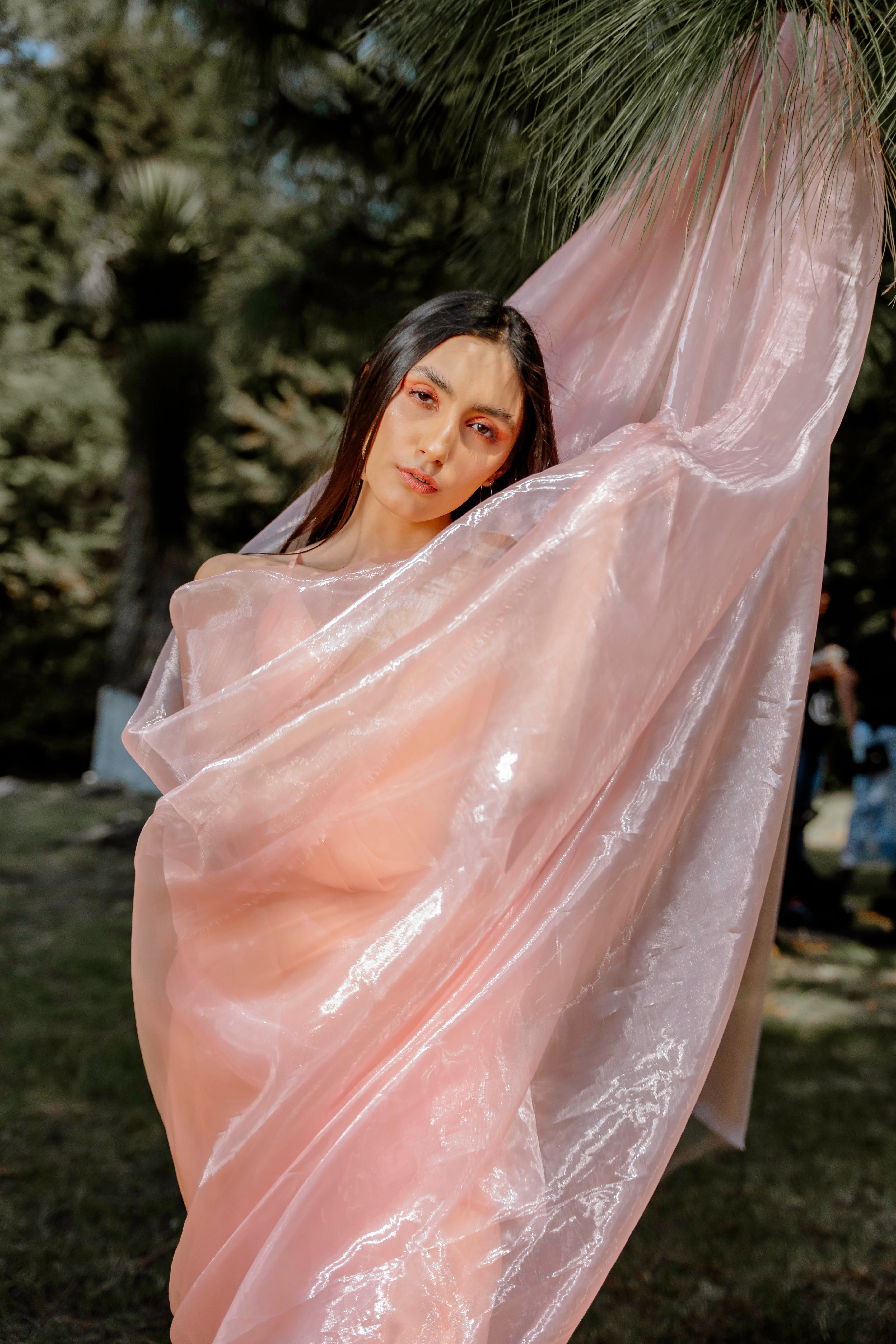 Elegant portrait of a woman draped in shimmering pink fabric, captured outdoors in a natural setting.