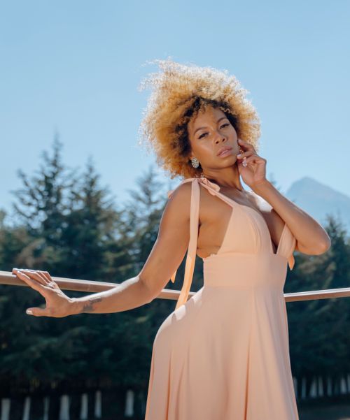 Free Fashion portrait of a woman in a peach dress posing outside with nature in the background. Stock Photo