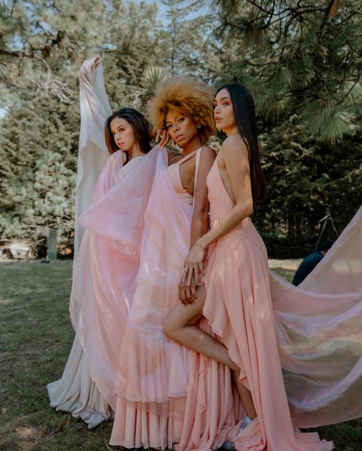 Three women elegantly pose in stylish pink dresses amidst lush greenery in a park.