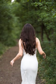 A woman in a white dress walks along a forest path surrounded by lush greenery.