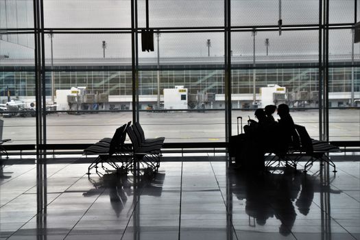 Silhouette of unrecognizable passengers sitting on chairs with luggage near  window and waiting for flight