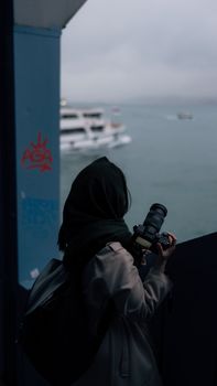A woman photographer captures a harbor view with her camera at dusk, showcasing the serene sea and moored boats.