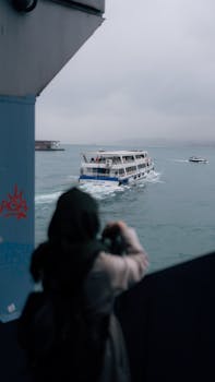 A woman in a hijab stands observing a passenger boat in overcast weather, creating a serene nautical scene.
