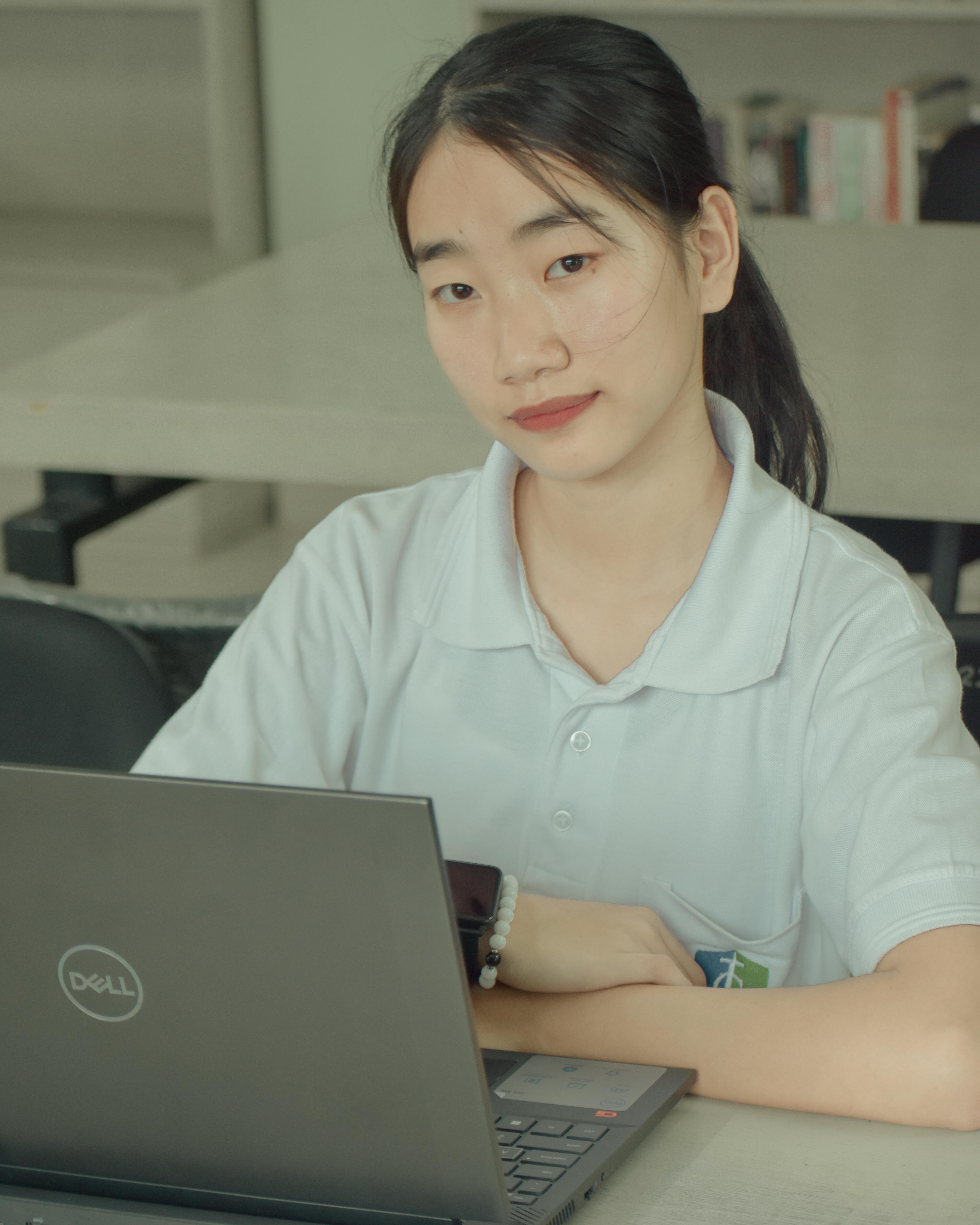 Asian female student sitting at a desk with a laptop, smiling indoors.