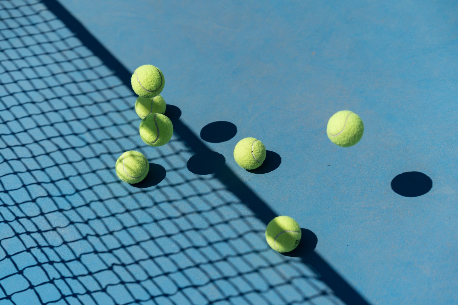 Tennis balls scattered on a blue hard court next to the net, bright sunlight and shadows