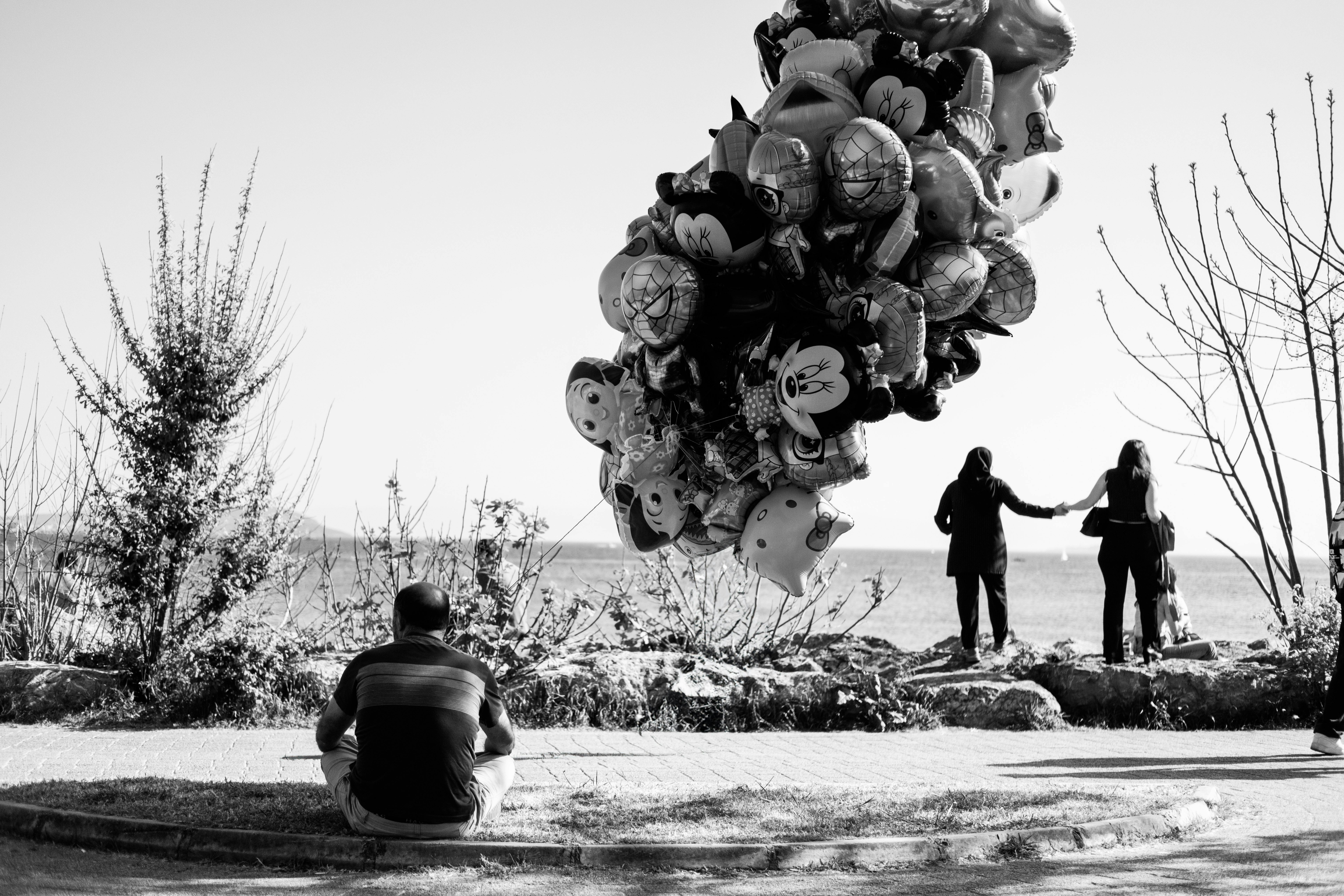 Man sitting with balloons overlooking a serene beach scene in black and white.