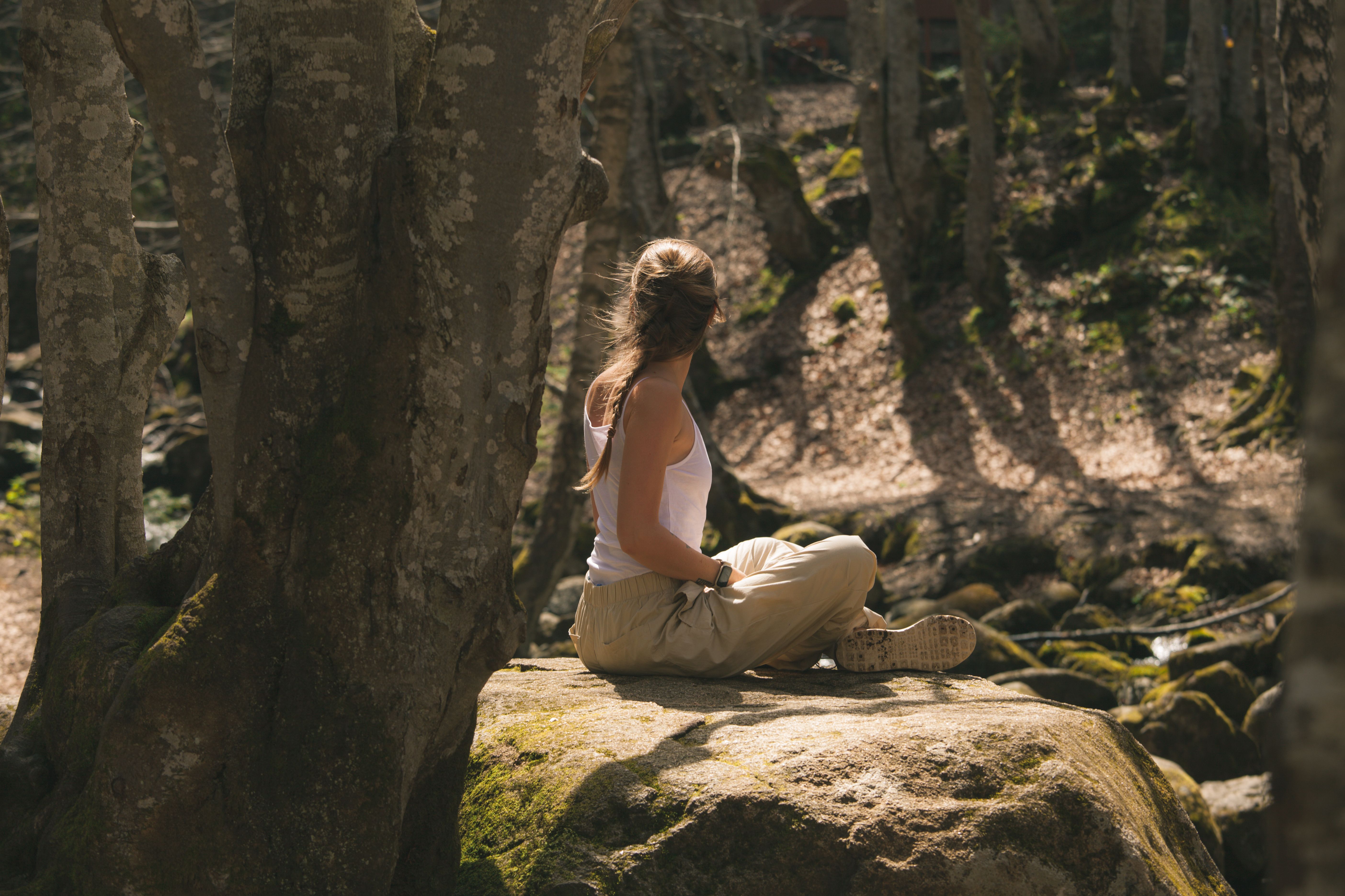 A woman in a white tank top enjoying solitude on a rock amidst a sun-dappled forest.