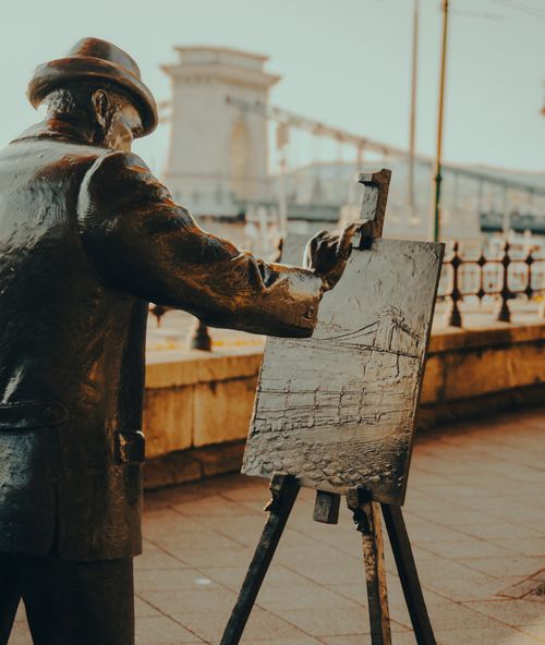 Free Bronze statue of a painter on a pavement with the Chain Bridge in Budapest, Hungary. Stock Photo