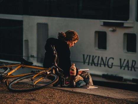 A serene moment of a woman reading beside her bicycle with a ship in the backdrop at sunset.