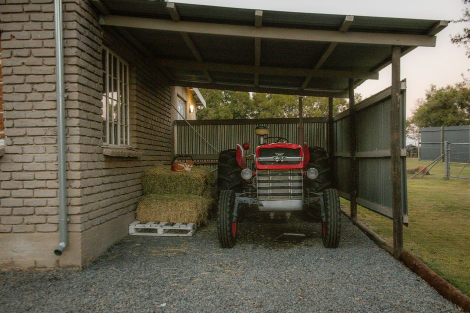 An old tractor parked under a rustic shed beside hay bales in a rural farm setting.