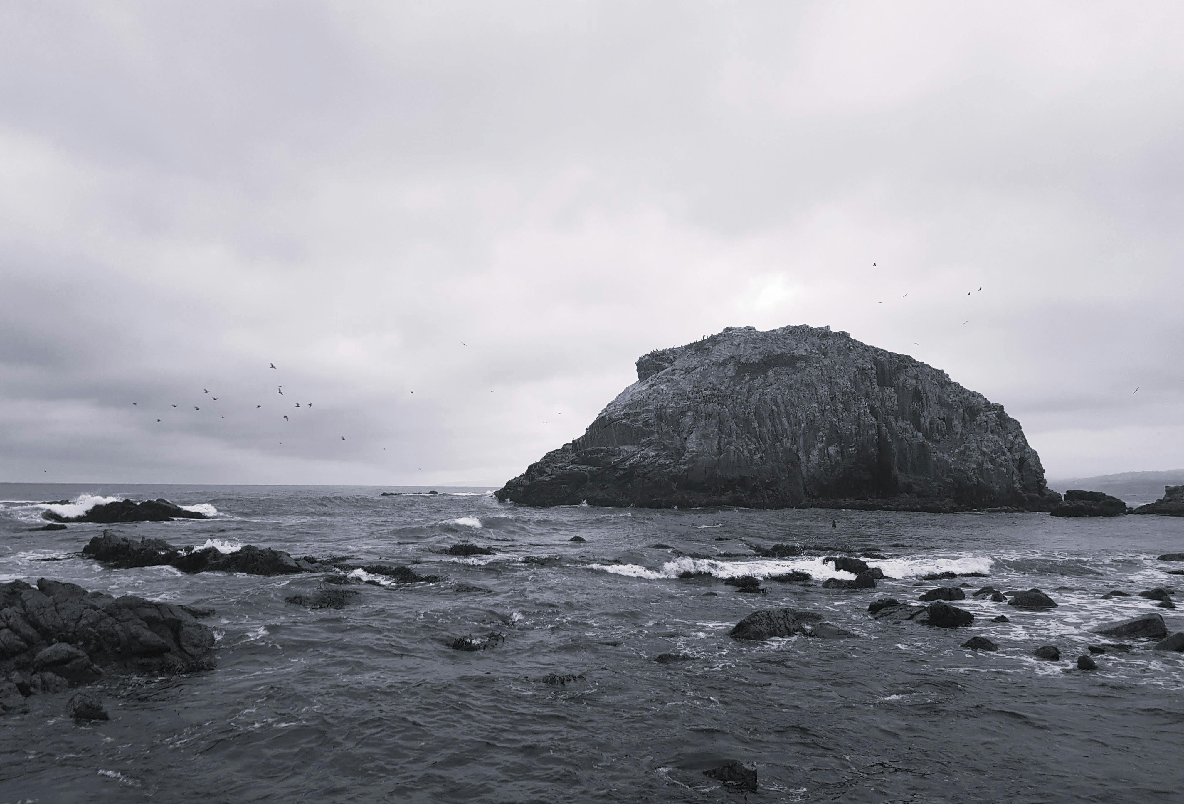 Moody seascape featuring a rocky coastline and island with a cloudy sky.