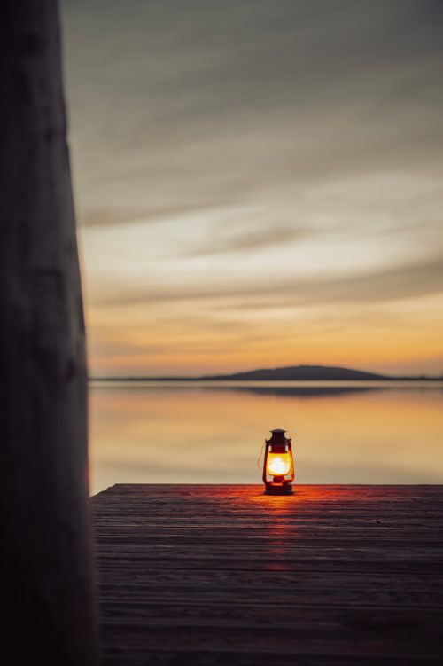 Free A tranquil lakeside scene at sunset in Leipzig with a lit kerosene lamp on a wooden pier, casting a warm glow. Stock Photo
