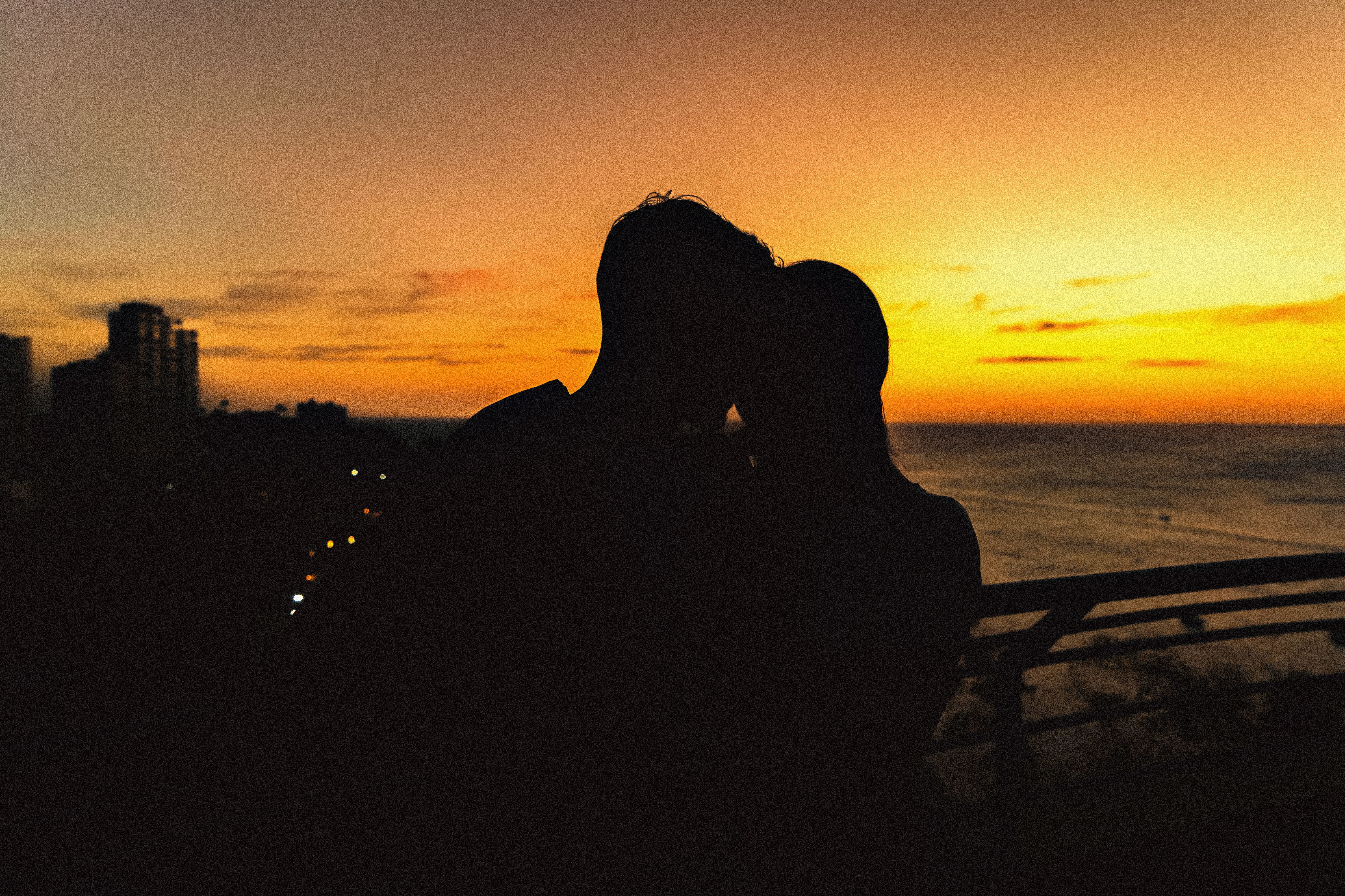 Silhouette of a couple sharing a romantic moment at sunset by the seaside with city lights in the background.