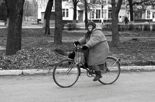 Black and white photo of a senior woman cycling in a city street during winter, wearing a headscarf and coat.