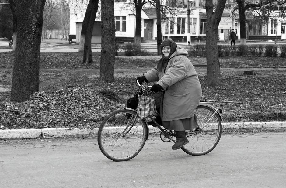 Black and white photo of a senior woman cycling in a city street during winter, wearing a headscarf and coat.