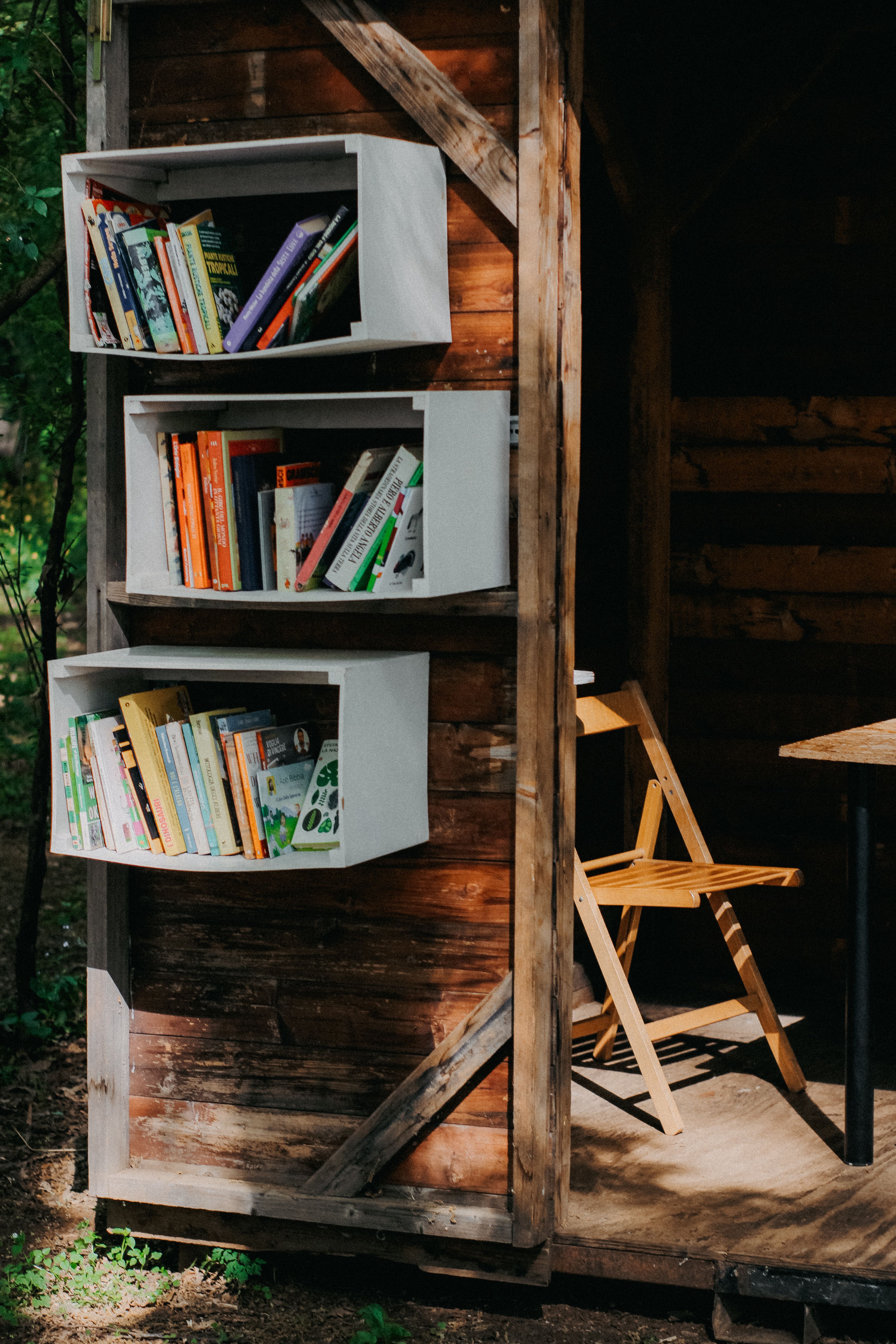 Wooden outdoor bookshelf with books and a folding chair, perfect for reading in nature.
