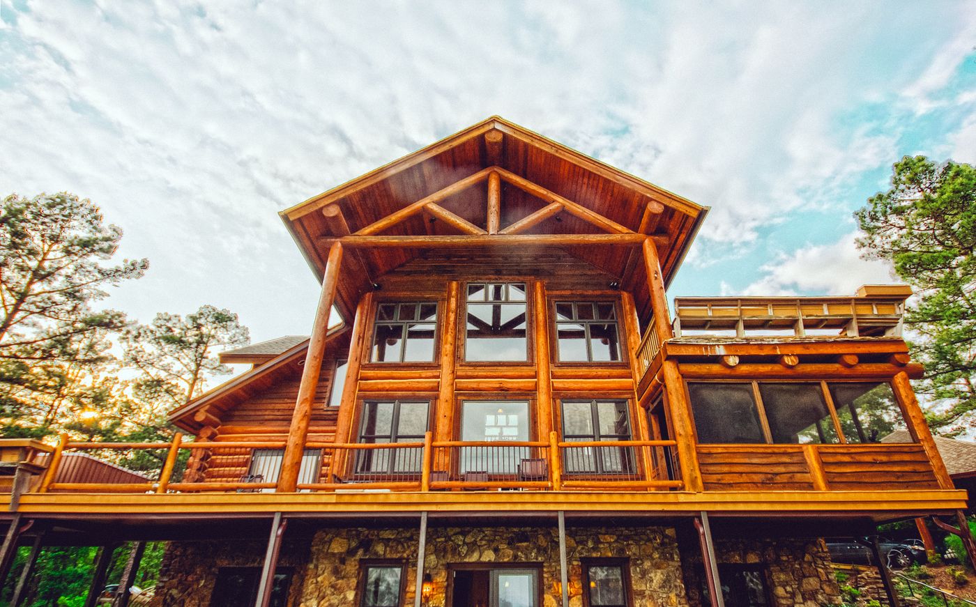 Large wood and stone cabin with tall windows and a deck framed by pine trees