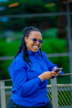 Fashionable woman in blue blouse smiling while holding a phone outdoors.