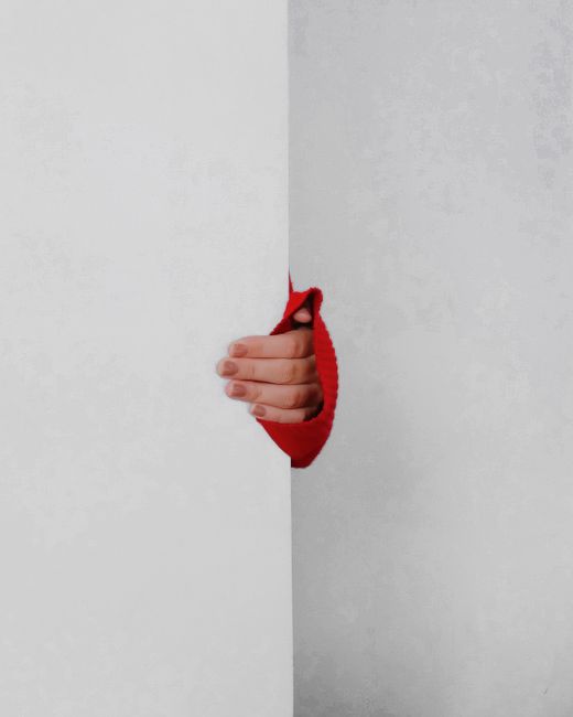 A person wears a red sweater and peeks a hand through a white wall, creating a minimalist and intriguing visual effect.