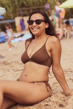 Woman in a bikini smiling on a sandy beach, enjoying a sunny summer day, wearing sunglasses.