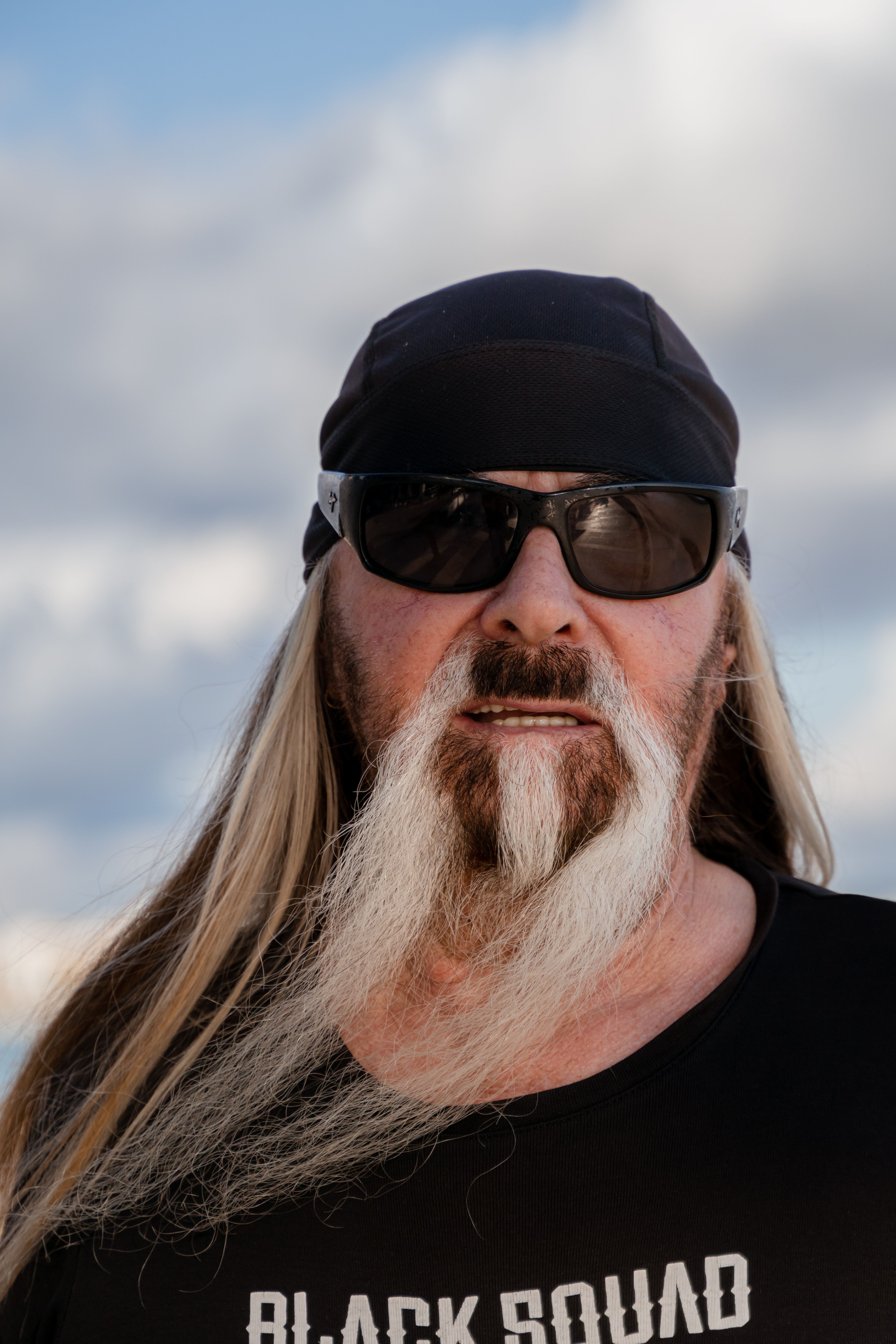 Portrait of a bearded man wearing sunglasses and a bandana, set against a cloudy sky.