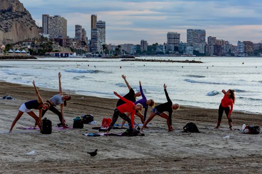 Group practicing yoga on Alicante beach with city skyline backdrop.