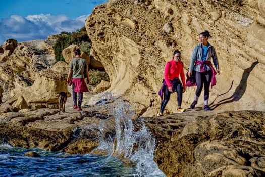 Hikers and a dog explore rocky coastal cliffs by the sea in Alicante, Spain.