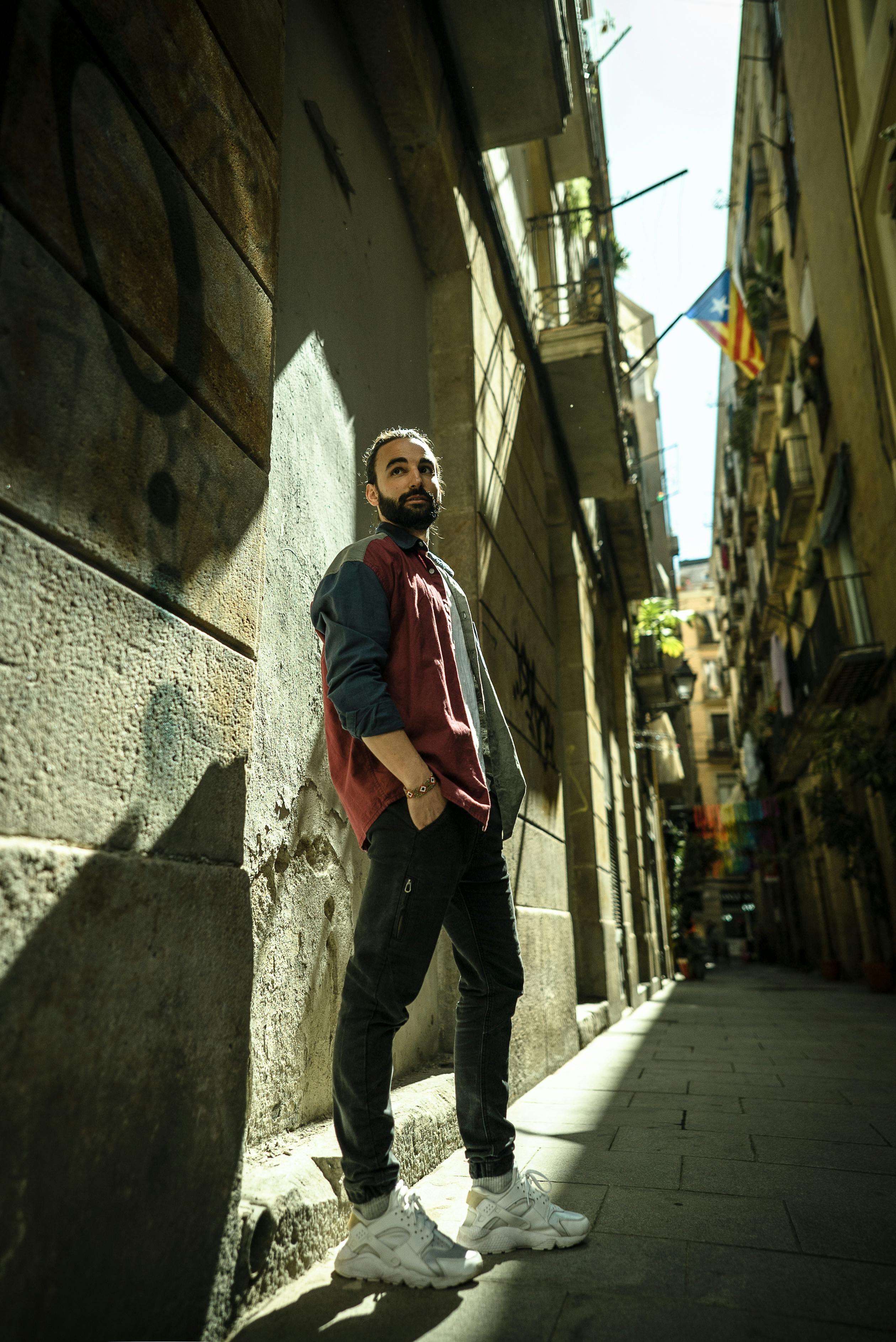 A fashionable man in red jacket poses in a sunlit narrow Barcelona alley.