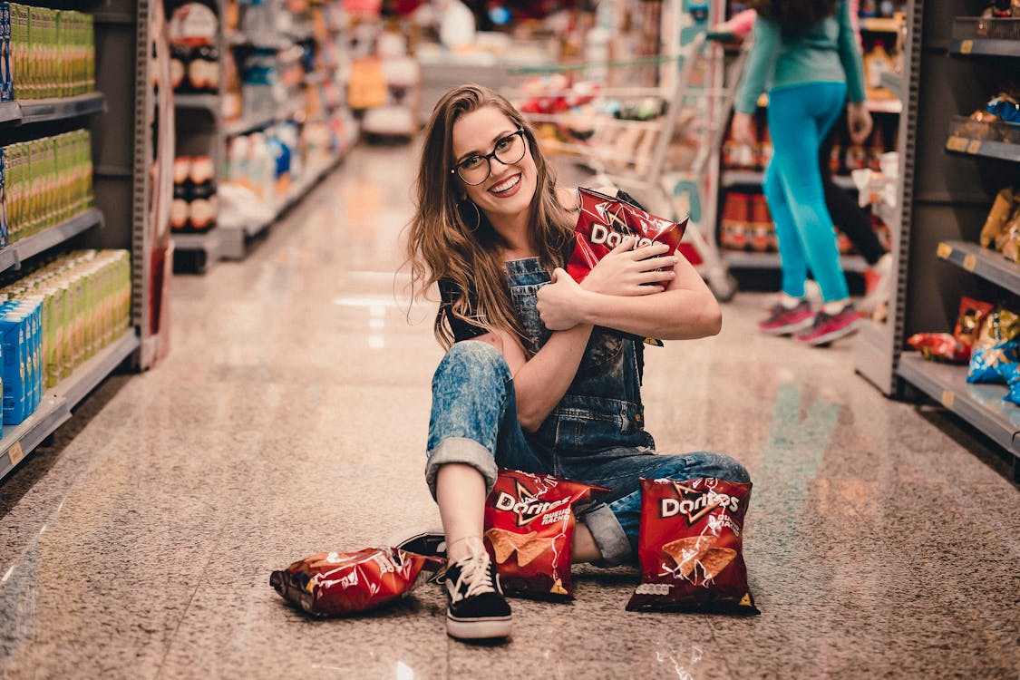Photo of Smiling Woman Sitting in the Middle of Shopping Aisle Holding Dorito Chips