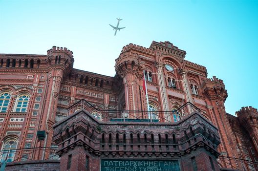 Low angle view of a historic red brick building with an airplane flying overhead in a clear blue sky.