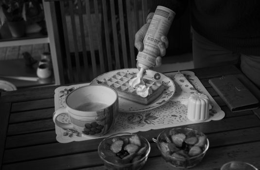 A close-up of a waffle with whipped cream, coffee, and fruit bowls in black and white.