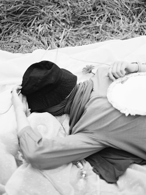 Black and White Photo of a Woman Lying on a Blanket with a Bucket Hat on Her Face