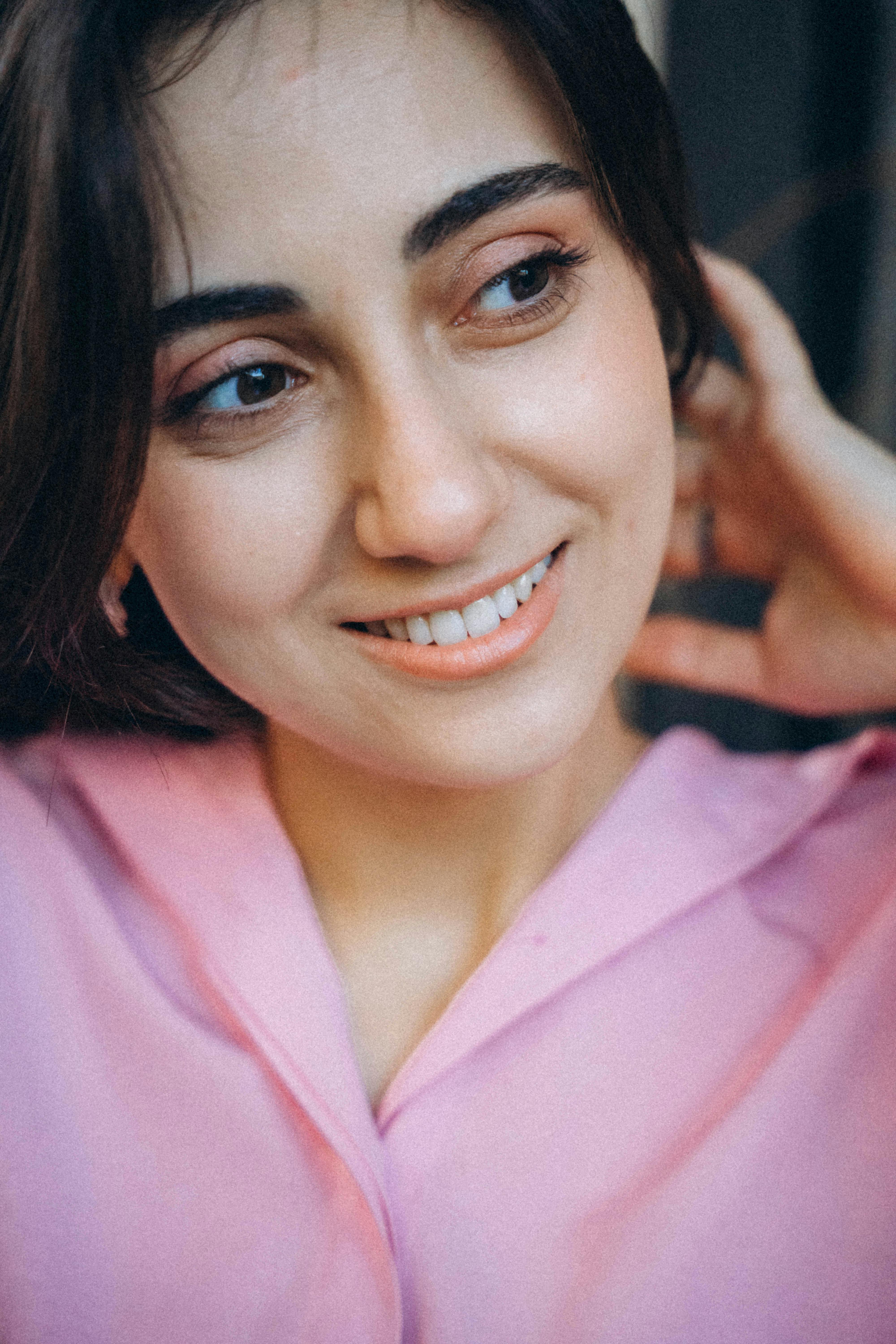 A cheerful woman with brunette hair in a pink shirt, smiling with a hand in her hair.